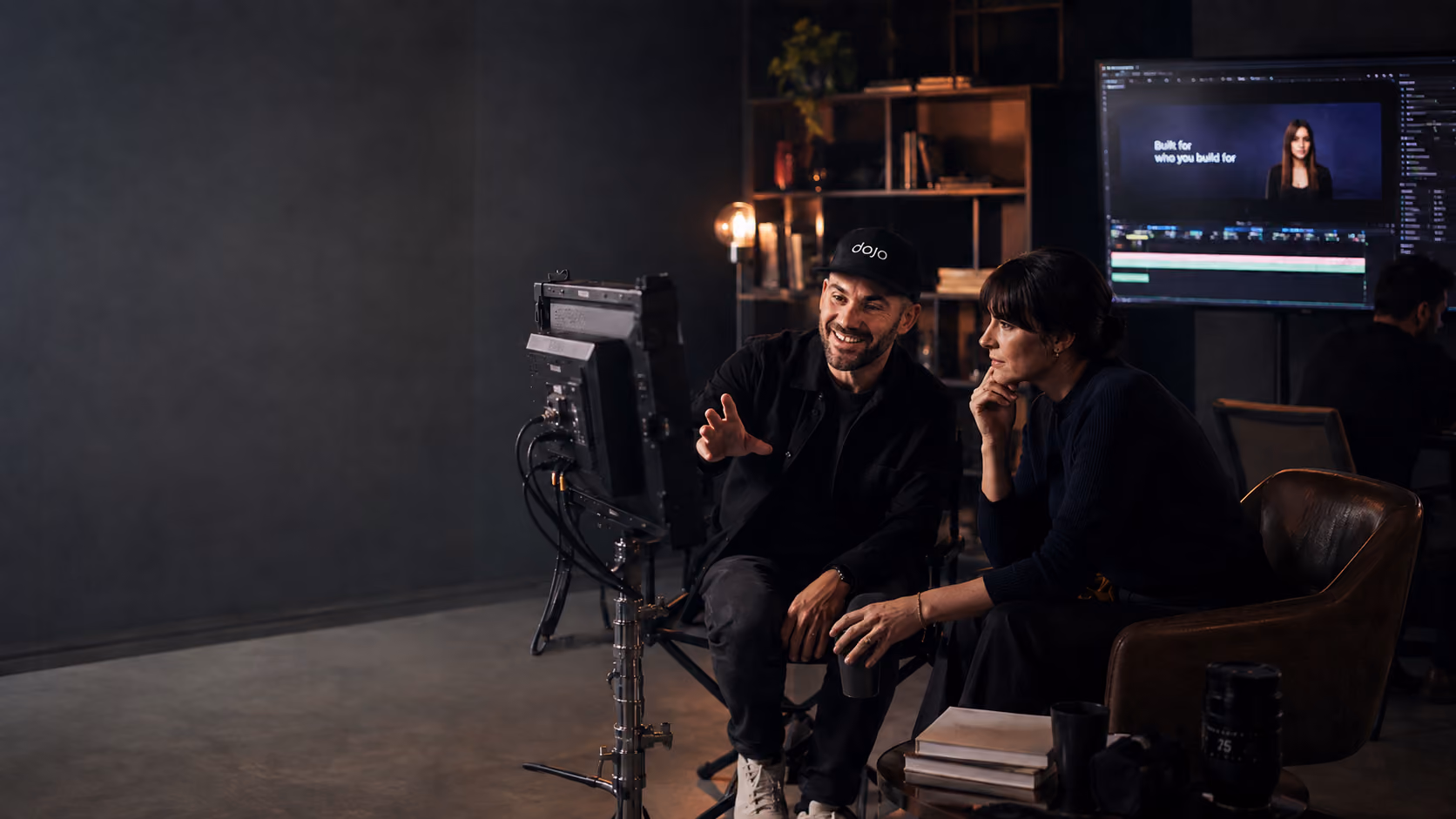 Two Dojo Films creatives reviewing a video edit together in a dark studio, with a monitor showing the footage timeline in the background