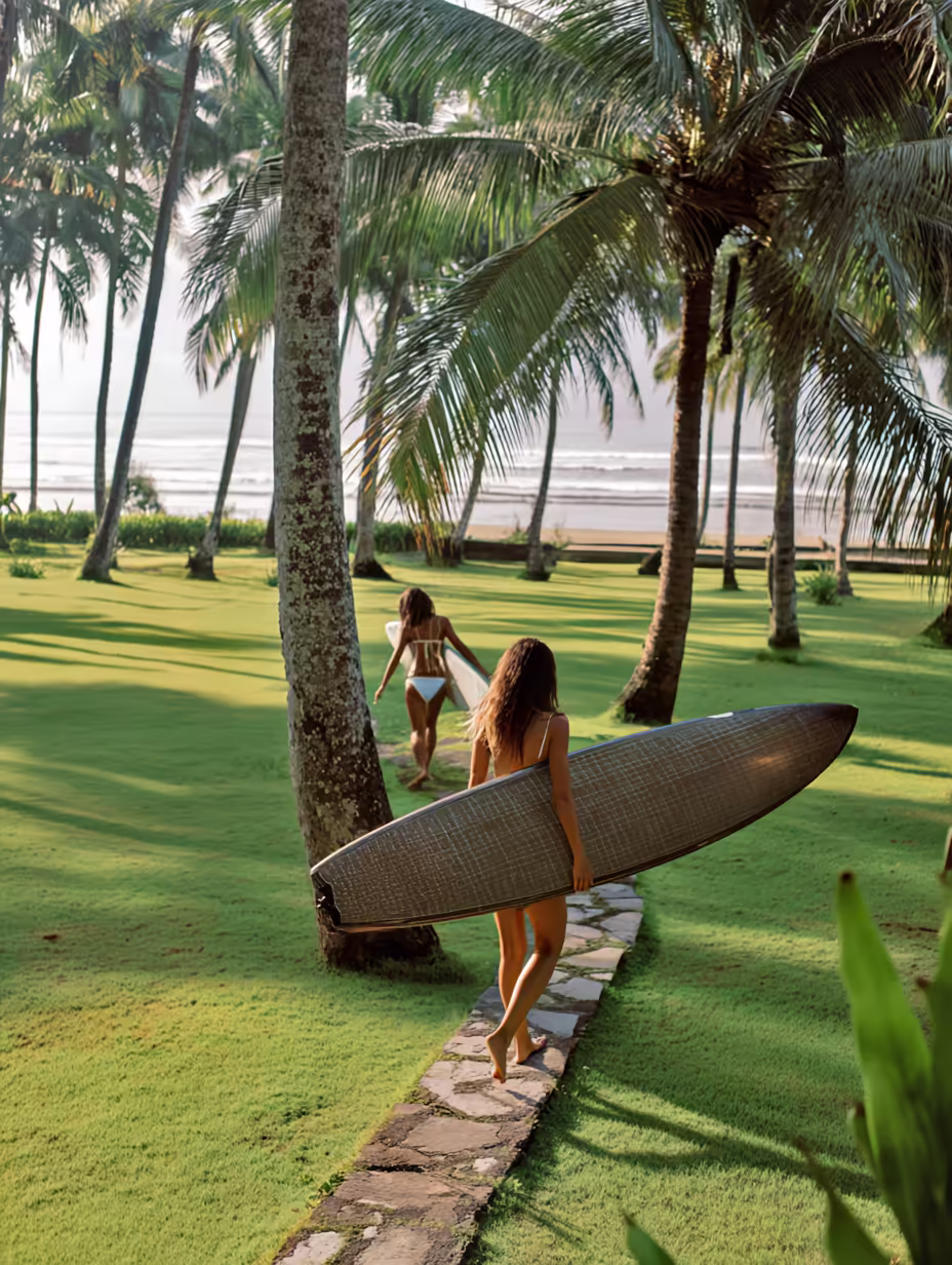 Two women in swimwear carrying surfboards while walking across a lush green lawn through a grove of palm trees toward the beach.