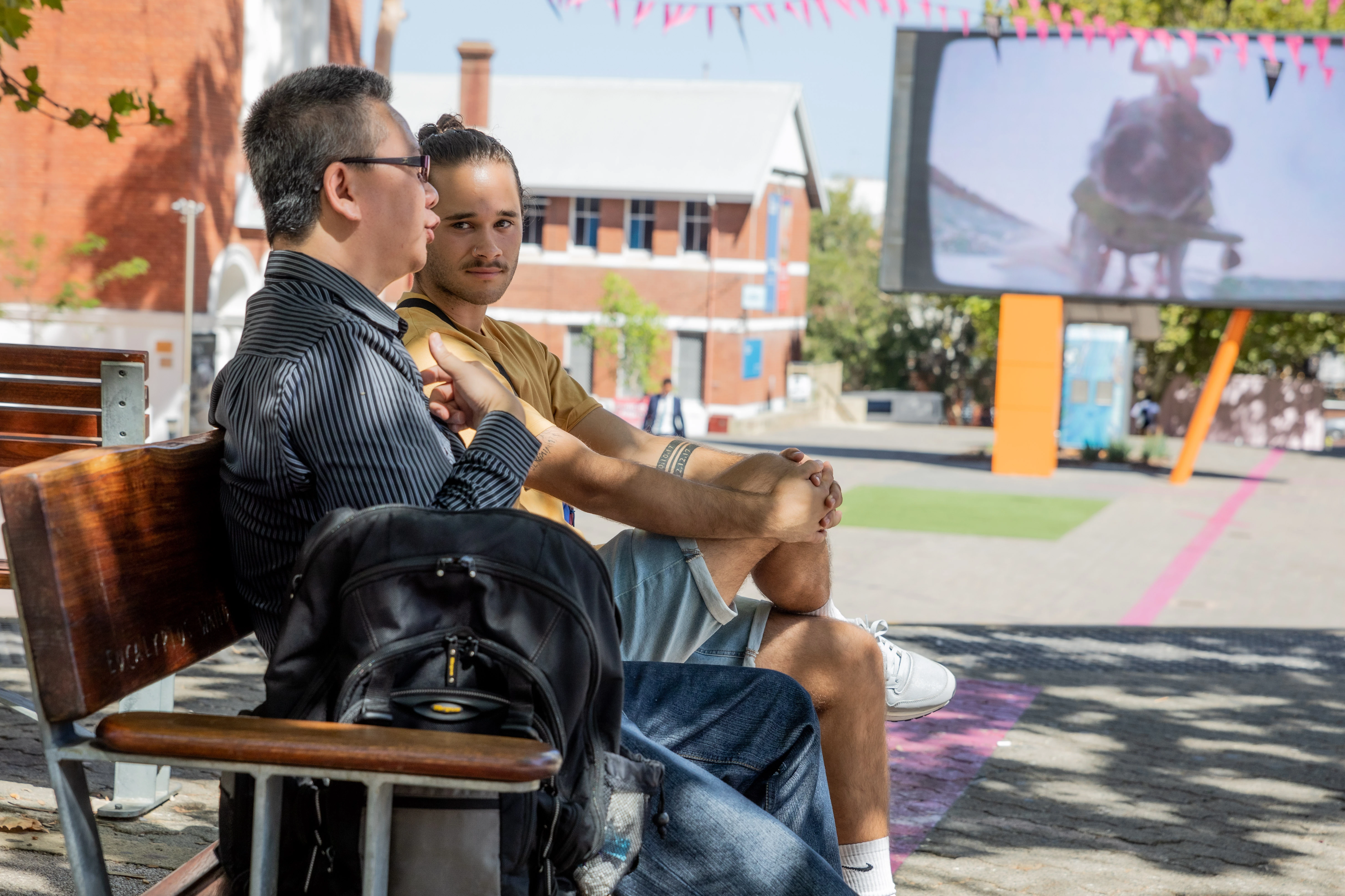 Two men sitting on a bench outdoors, one talking and the other listening attentively.
