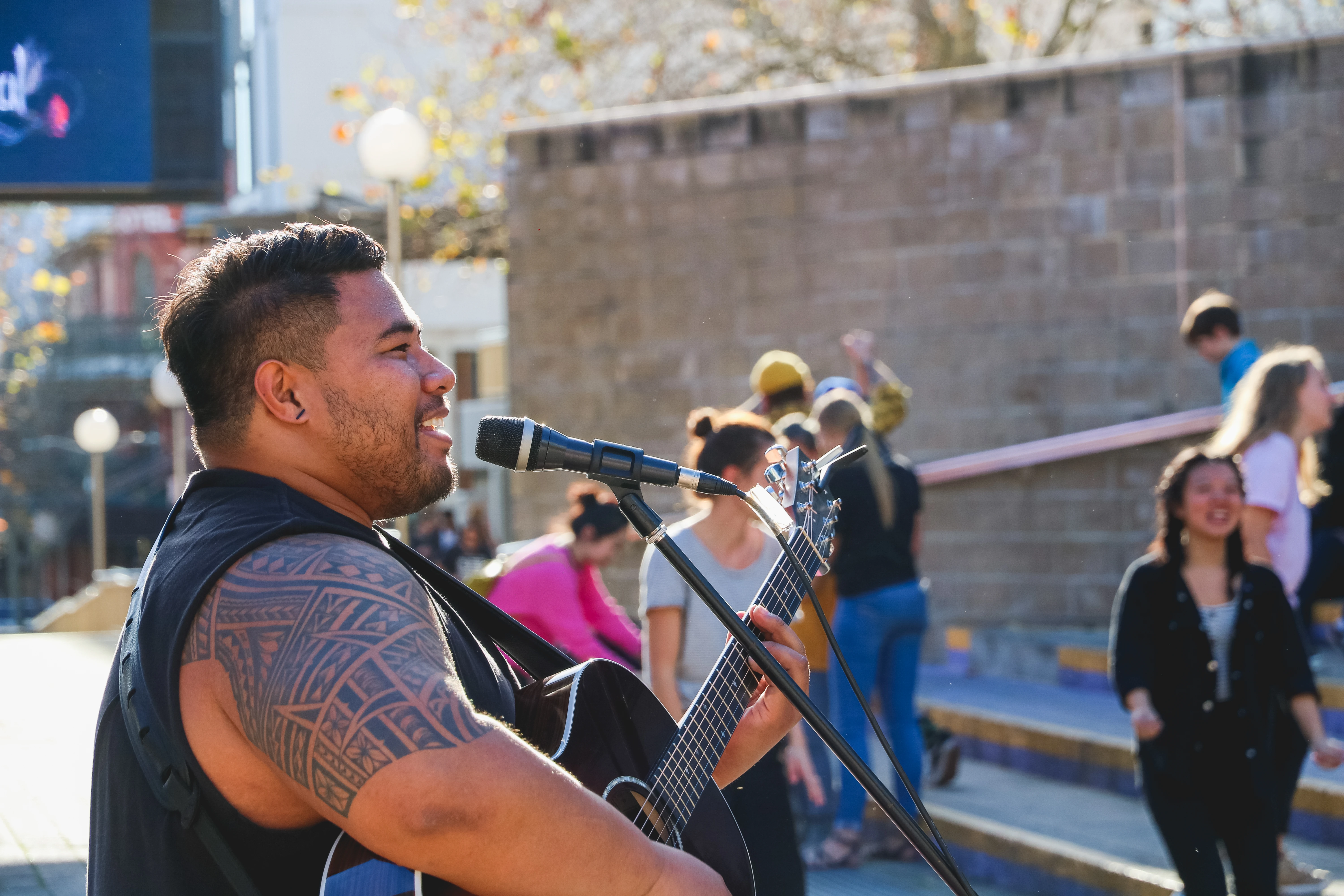Man with a tribal tattoo playing guitar and singing into a microphone outdoors during daytime.