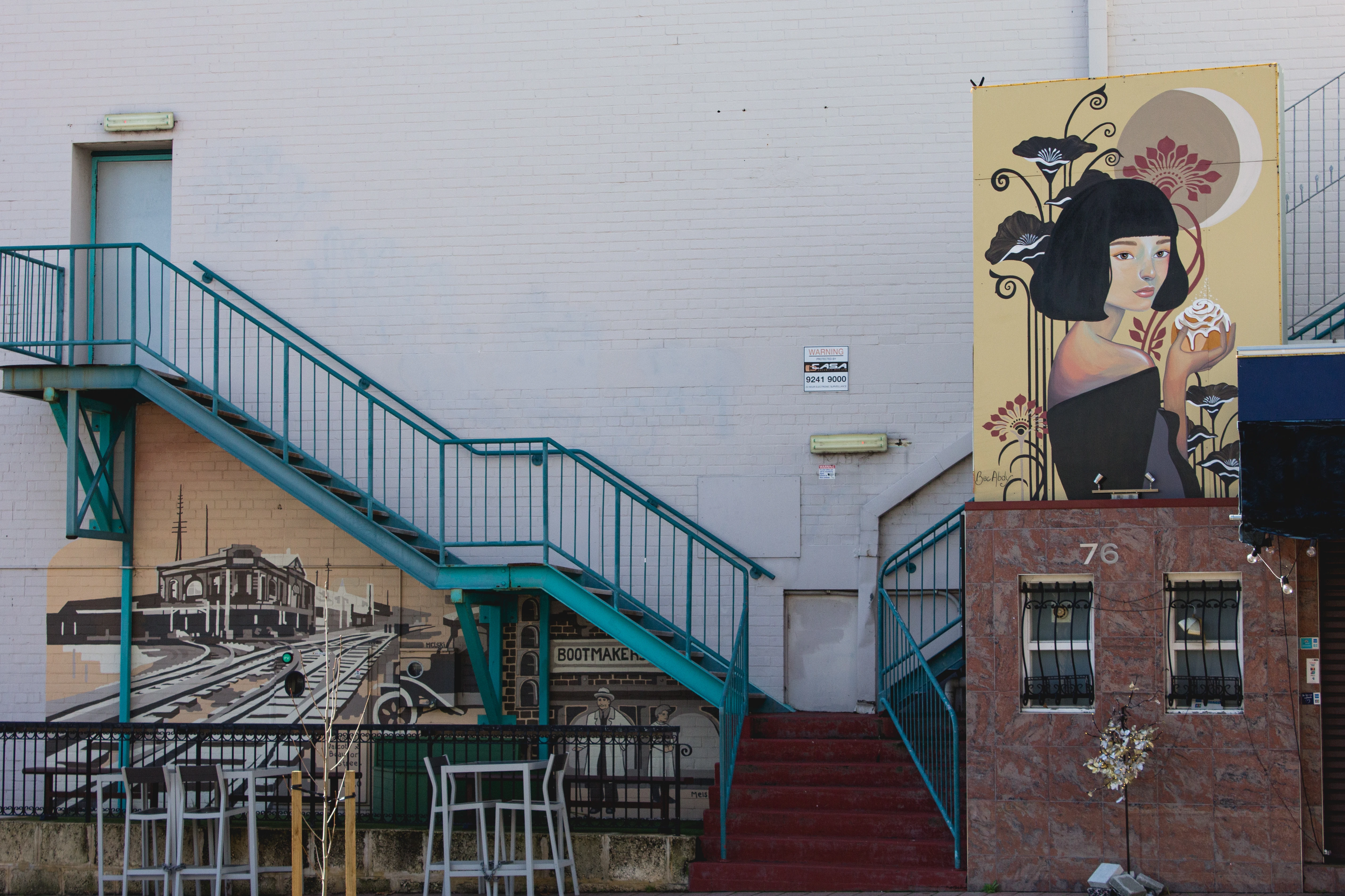 Outdoor stairs with teal railing by a wall featuring a mural of a woman holding a pastry and vintage street scene.