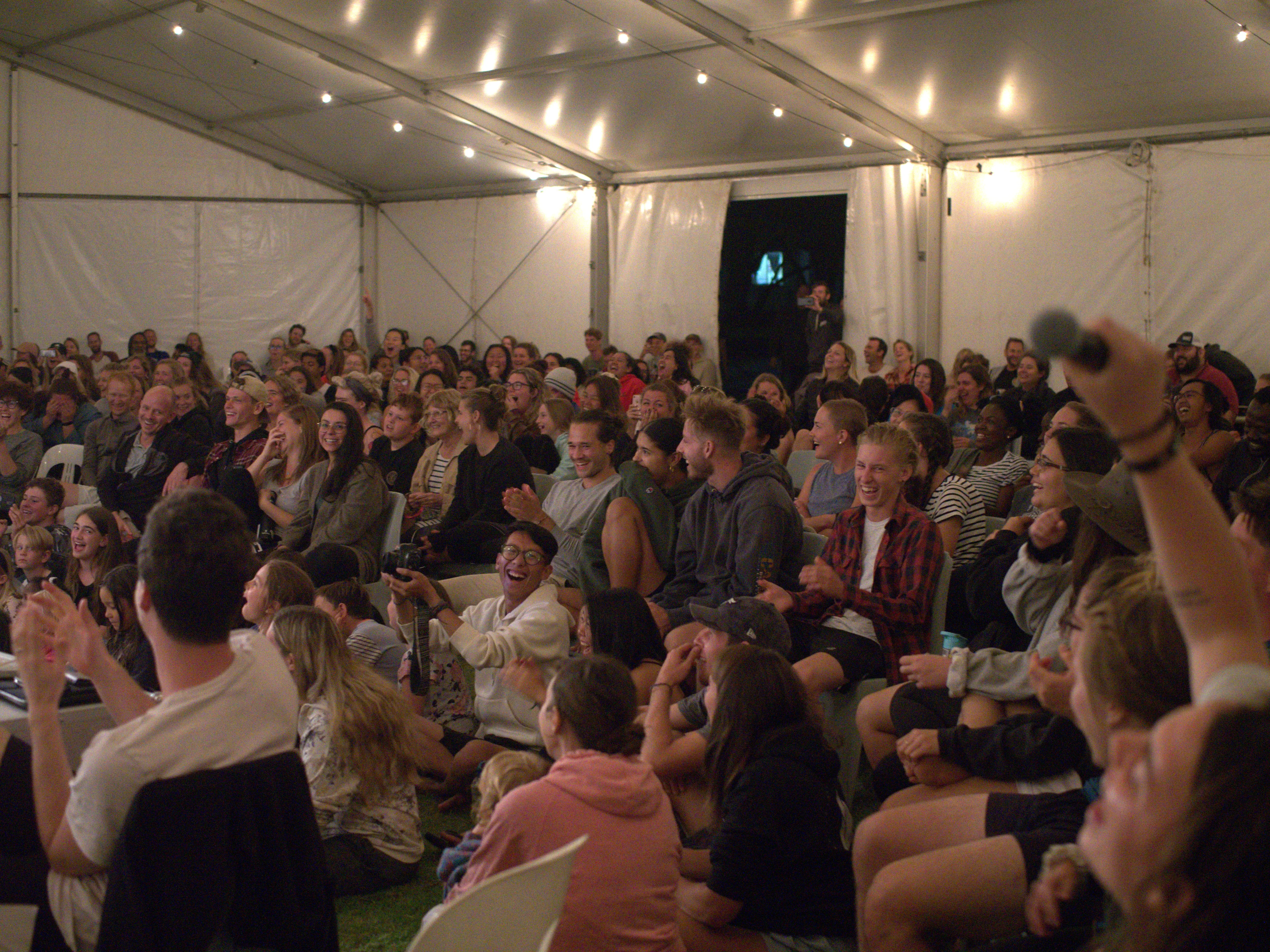 A large, diverse audience seated in a tent, watching and laughing, with a hand holding a microphone visible.