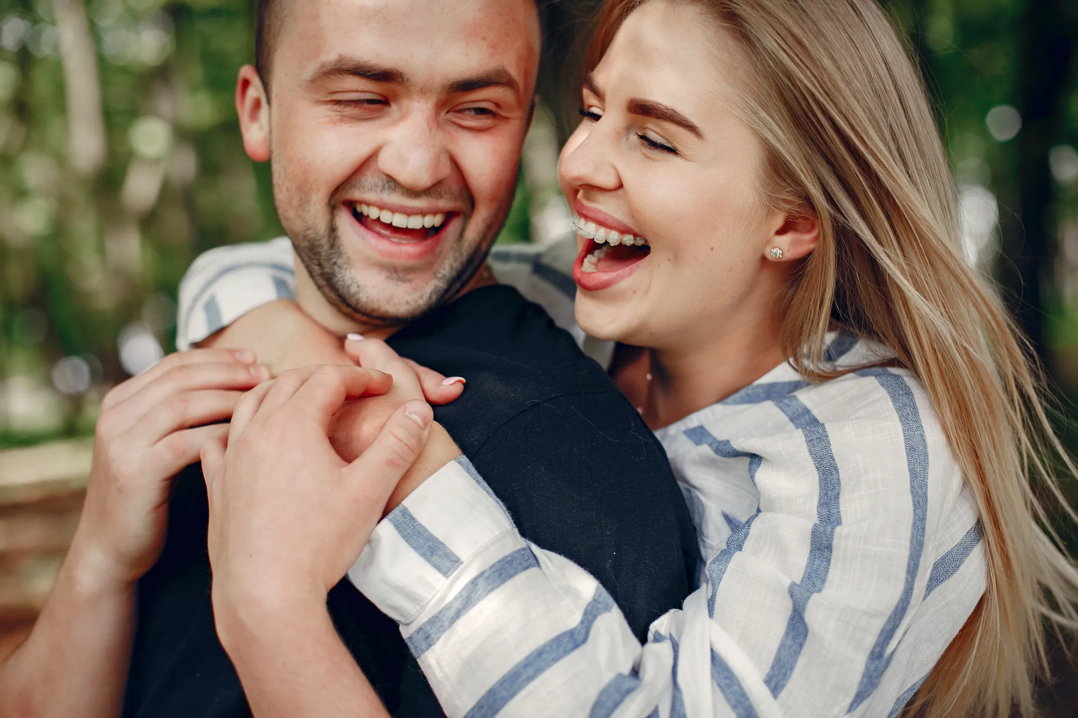 Smiling woman with braces hugging happy man from behind outdoors with blurred green background.