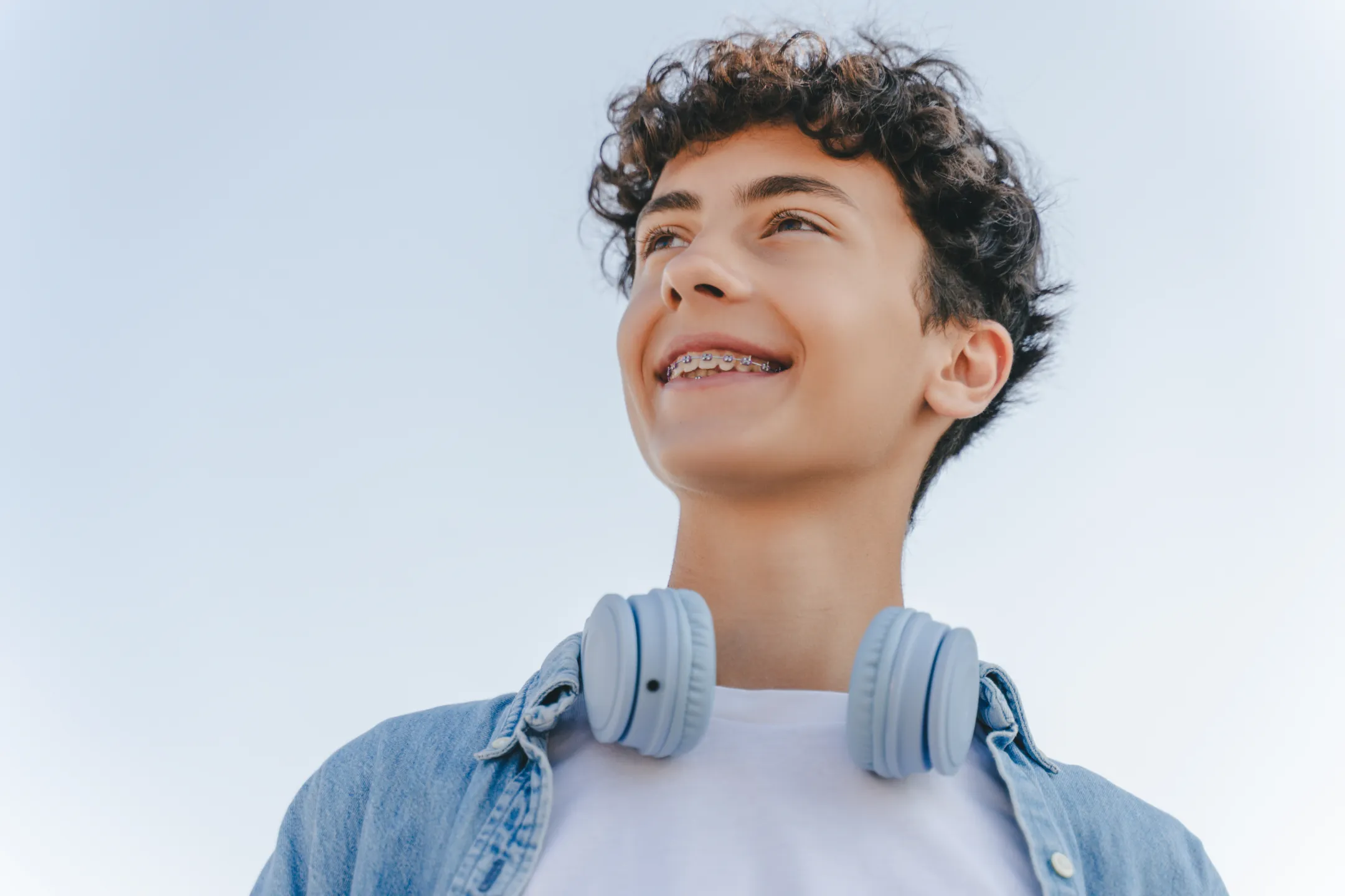 Teenage boy with curly hair and braces smiling, wearing headphones and denim jacket under clear sky.
