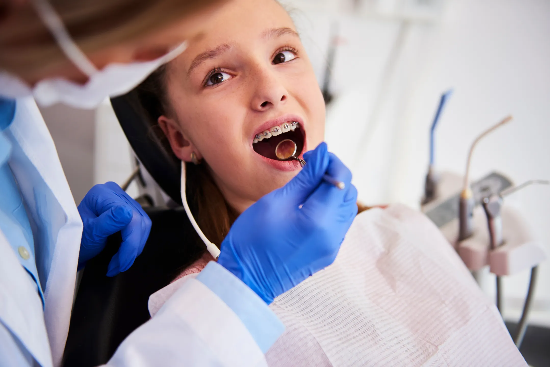 Girl with braces in dental chair having her teeth examined by dentist with blue gloves.