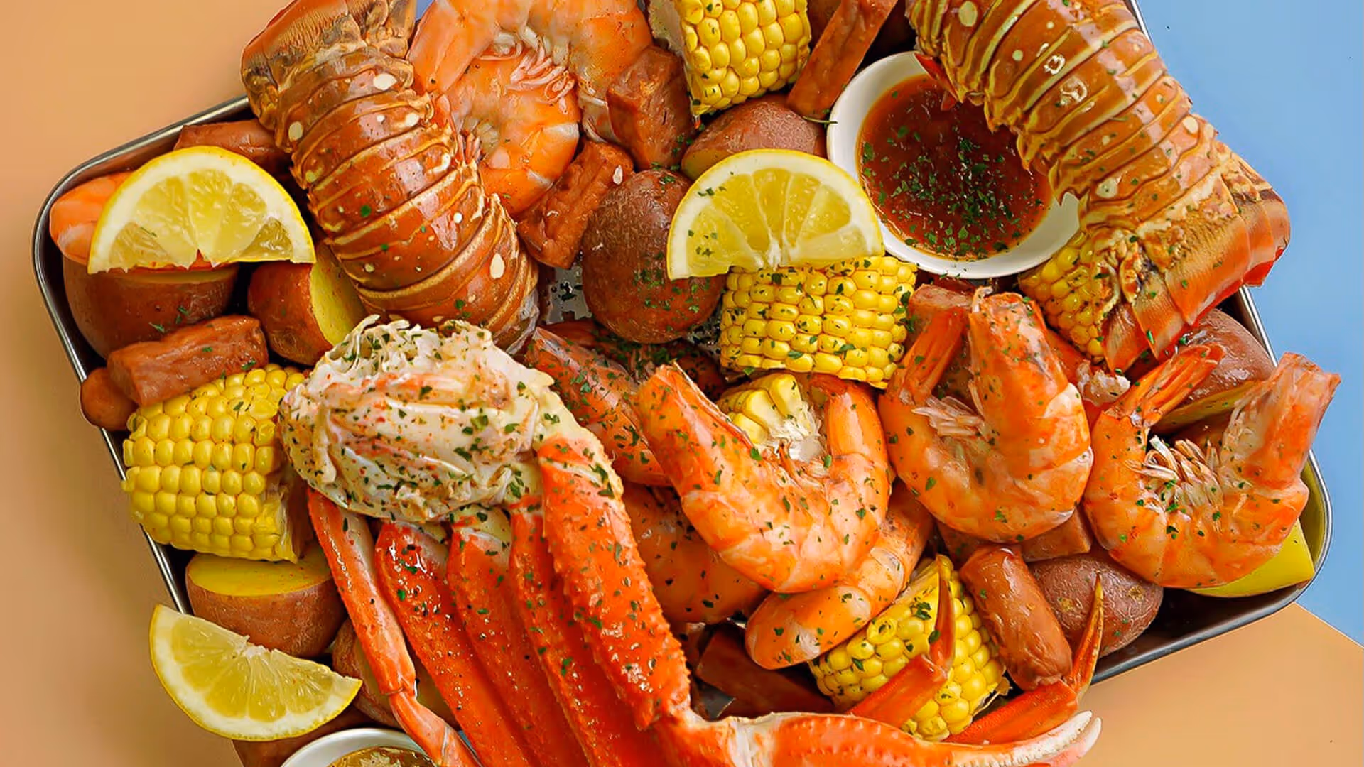 Seafood boil platter with lobster tails, crab legs, shrimp, corn, potatoes, sausage, lemon wedges, and dipping sauce.