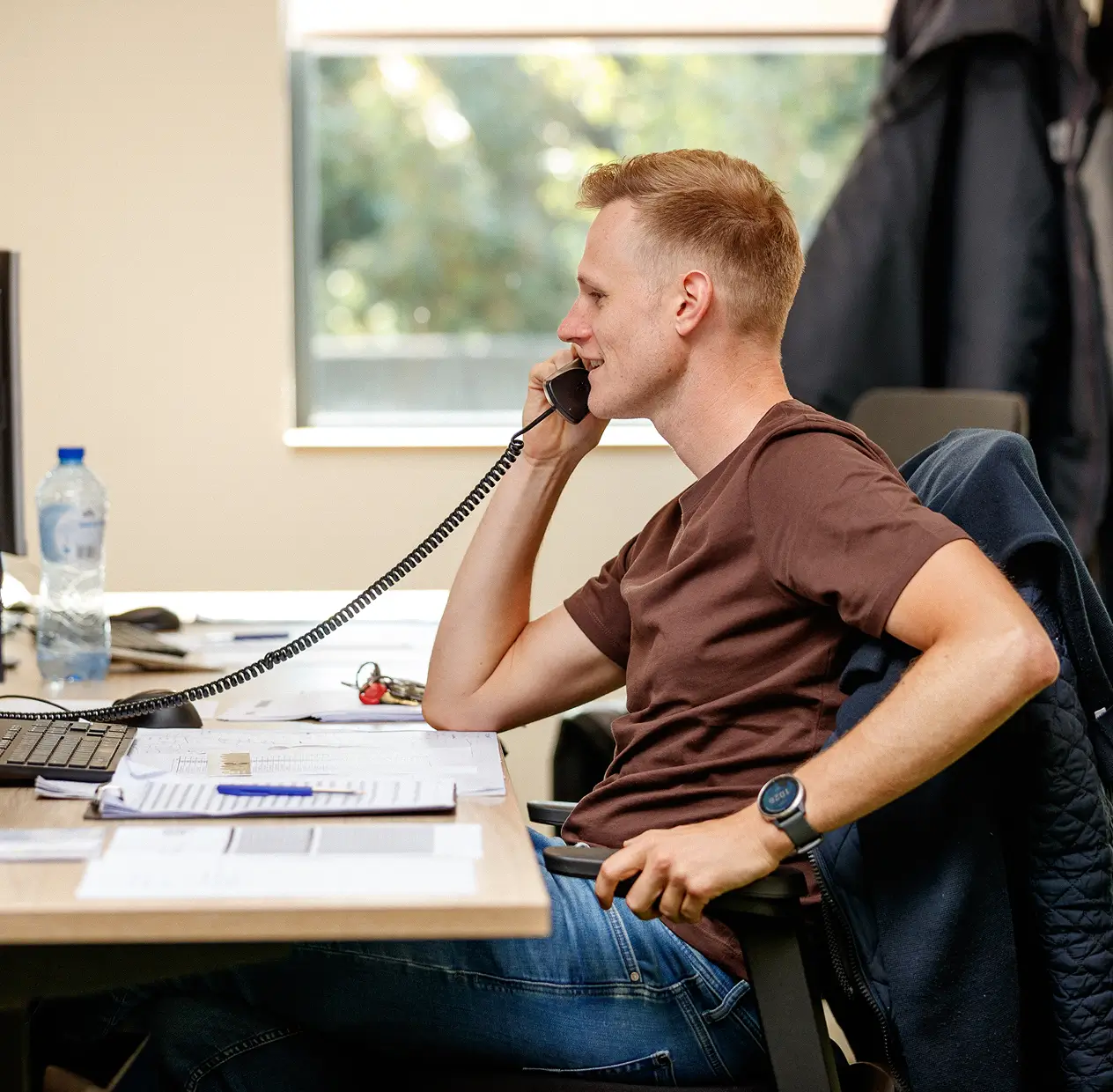 Man in shirt and jeans sitting at desk talking on a corded phone in an office.