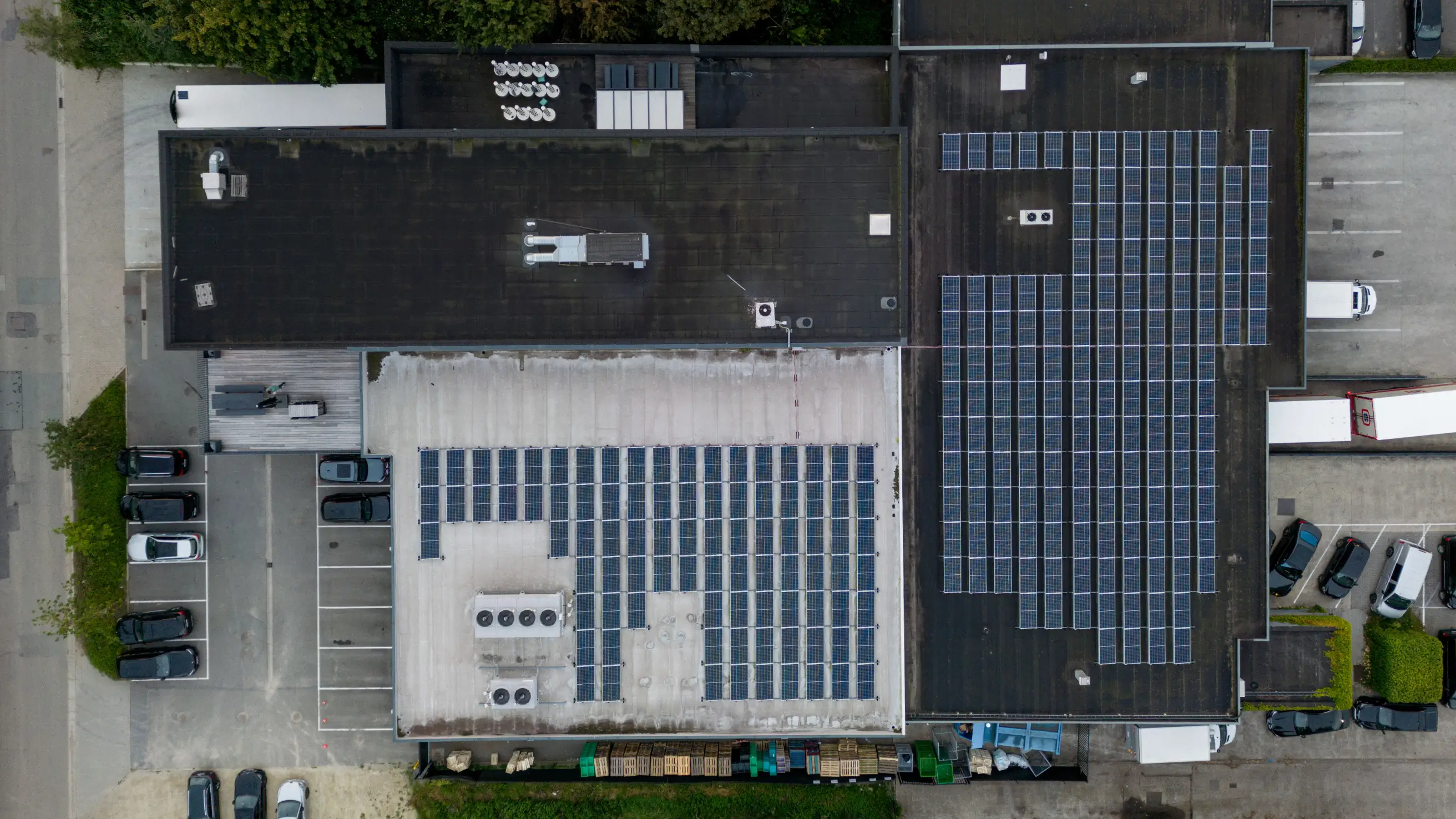 Luchtfoto van bedrijfsdaken met zonnepanelen en geparkeerde auto's rondom de gebouwen.