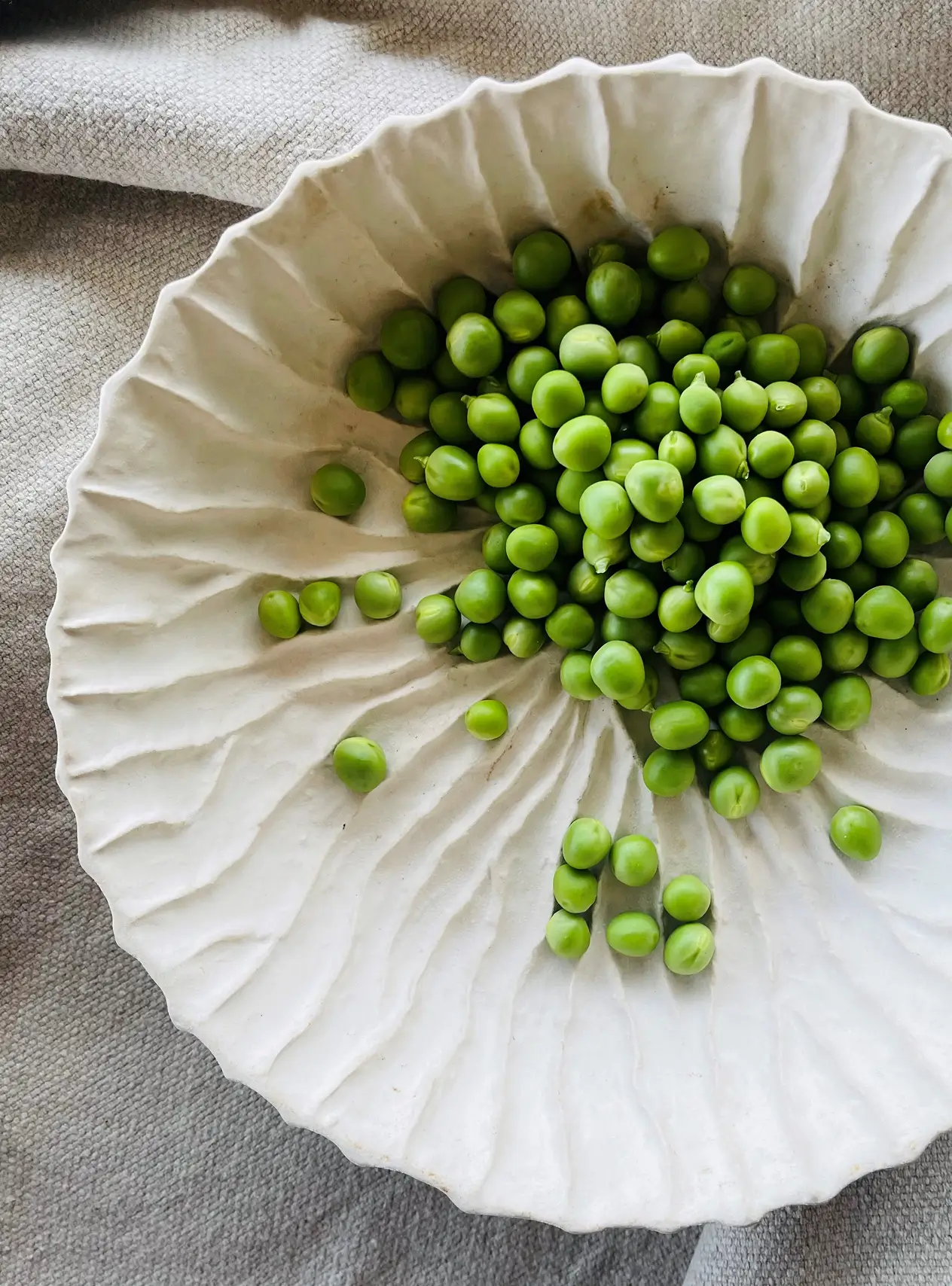 Witte bord met groene doperwten erop, geplaatst op een grijze stoffen ondergrond.