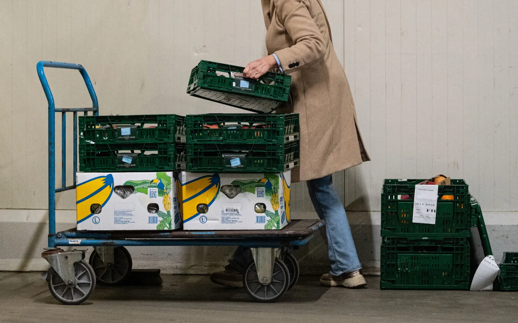 Persoon in lange jas plaatst groene kratjes op een blauwe steekwagen met dozen.