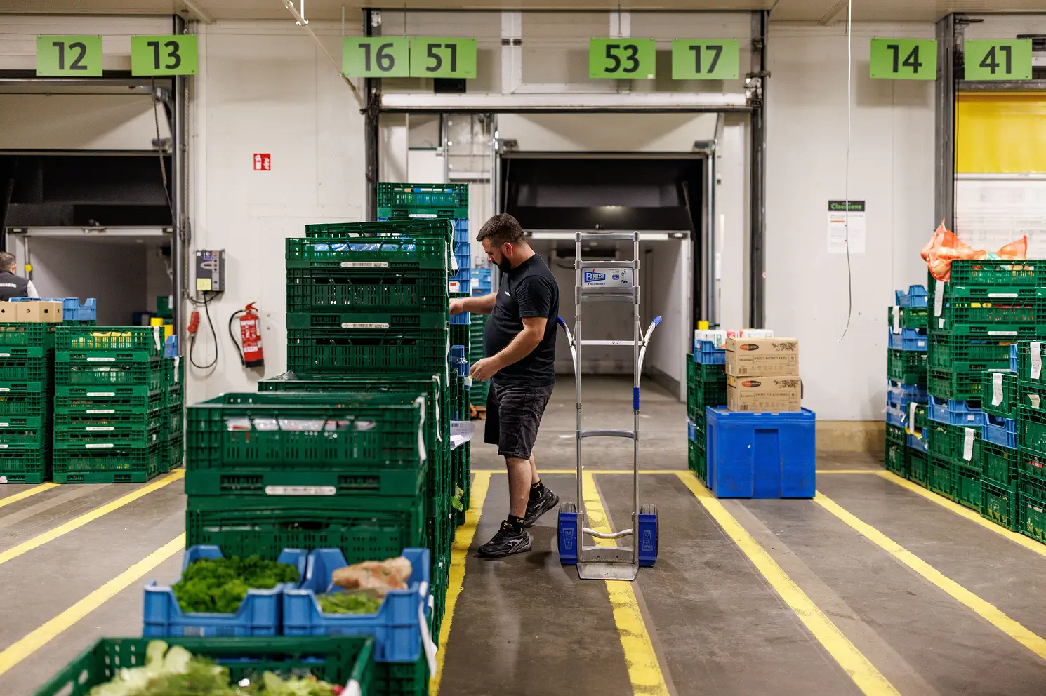 Man werkt in een magazijn met stapels groene kratten en een leeg steekwagentje naast hem.