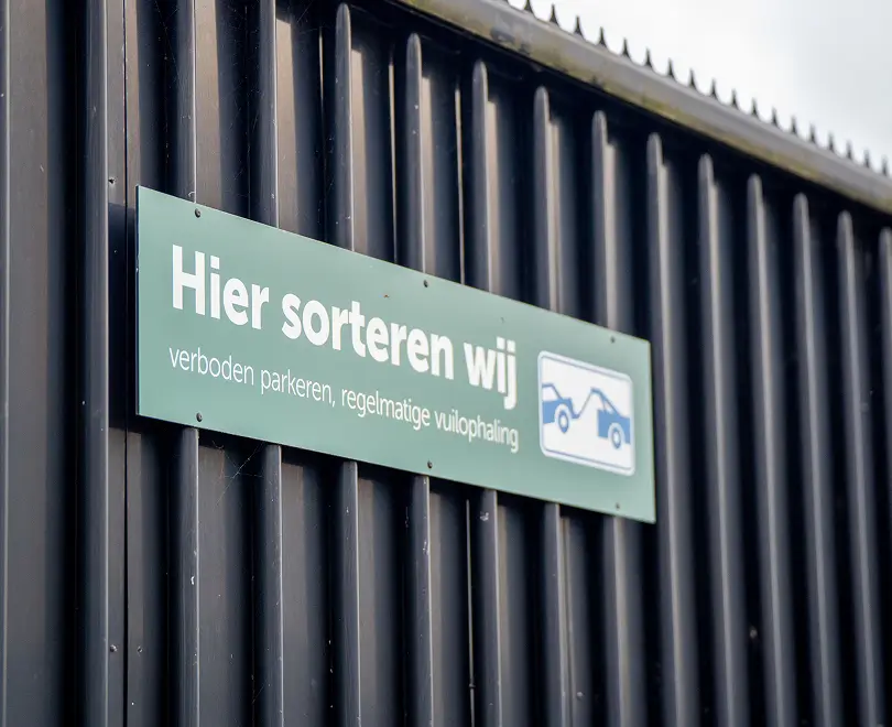 Groen bord aan zwarte muur met tekst 'Hier sorteren wij, verboden parkeren, regelmatige vuilophaling'.