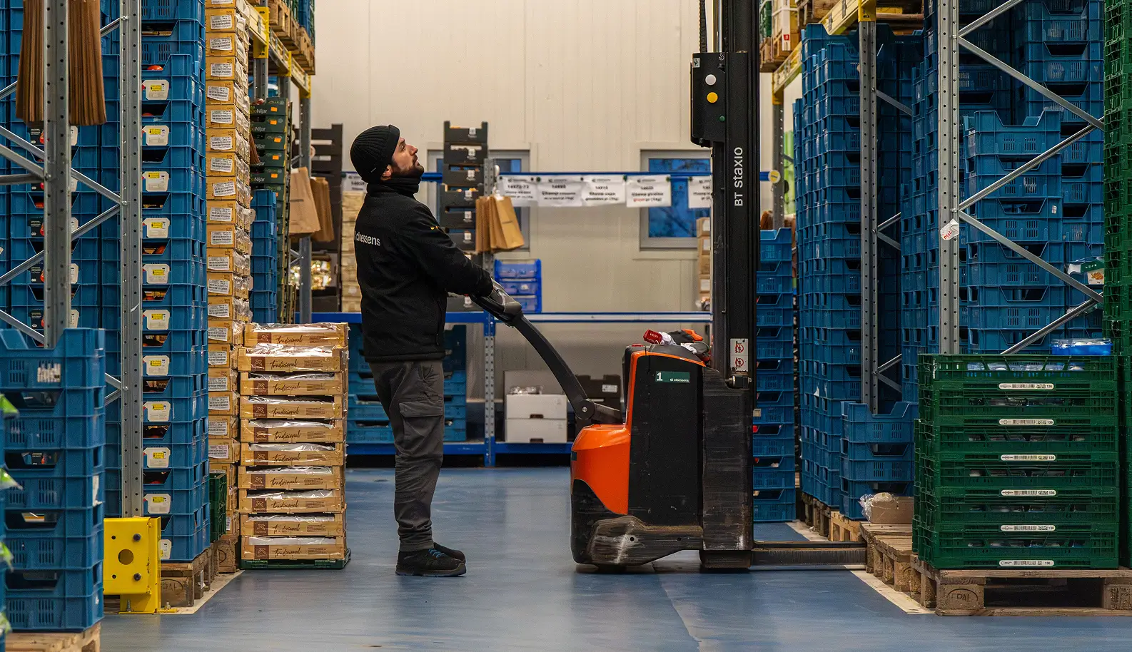 Werknemer bedient een palletwagen in een magazijn tussen gestapelde dozen en kratten.