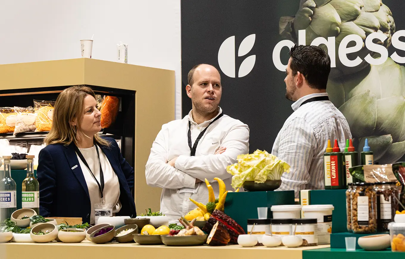 Drie personen in gesprek bij een stand met vers voedsel en producten tijdens een beurs.