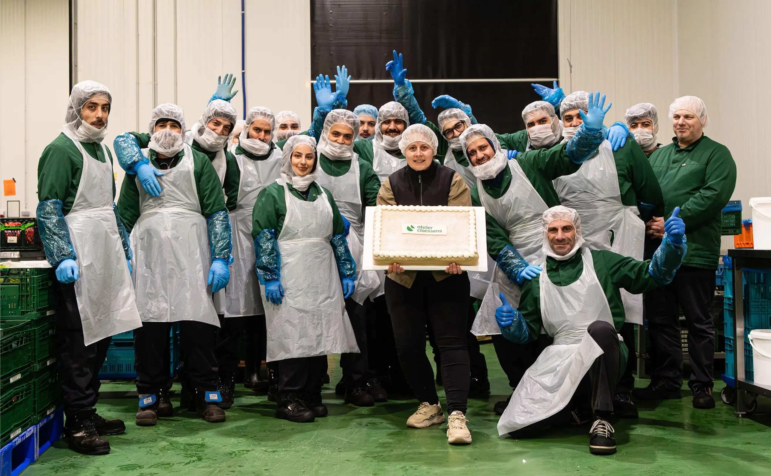 Team in protective gear posing indoors, one person holding a cake with the Atelier Claessens logo.