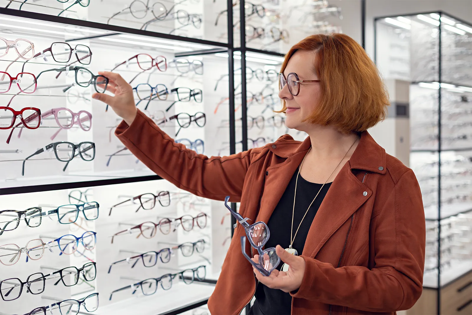 Woman with red hair trying on eyeglasses at an optical store display stand.