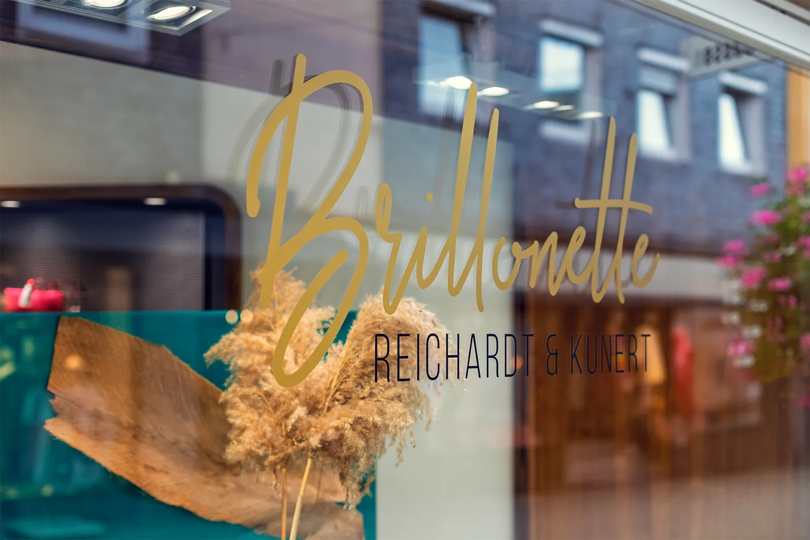 Shop window with gold script text reading 'Brillonette Reichardt & Kunert' and dried plant decor inside.
