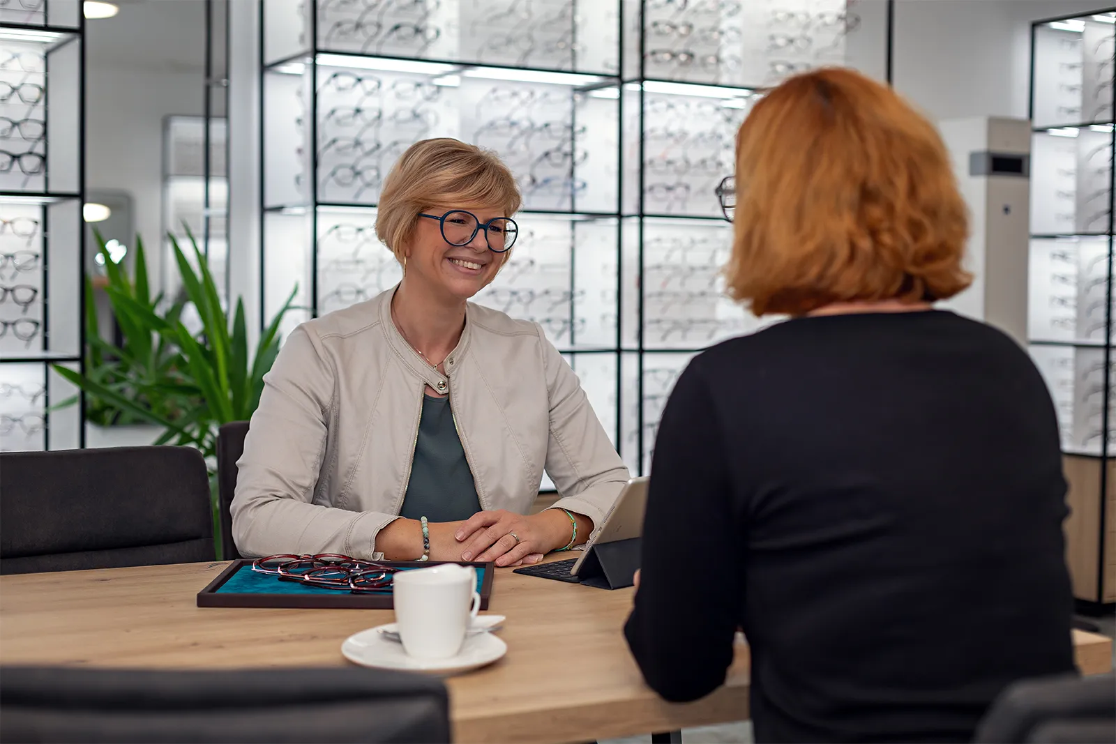Optician in glasses smiling and consulting a customer at a desk with multiple eyeglass frames.