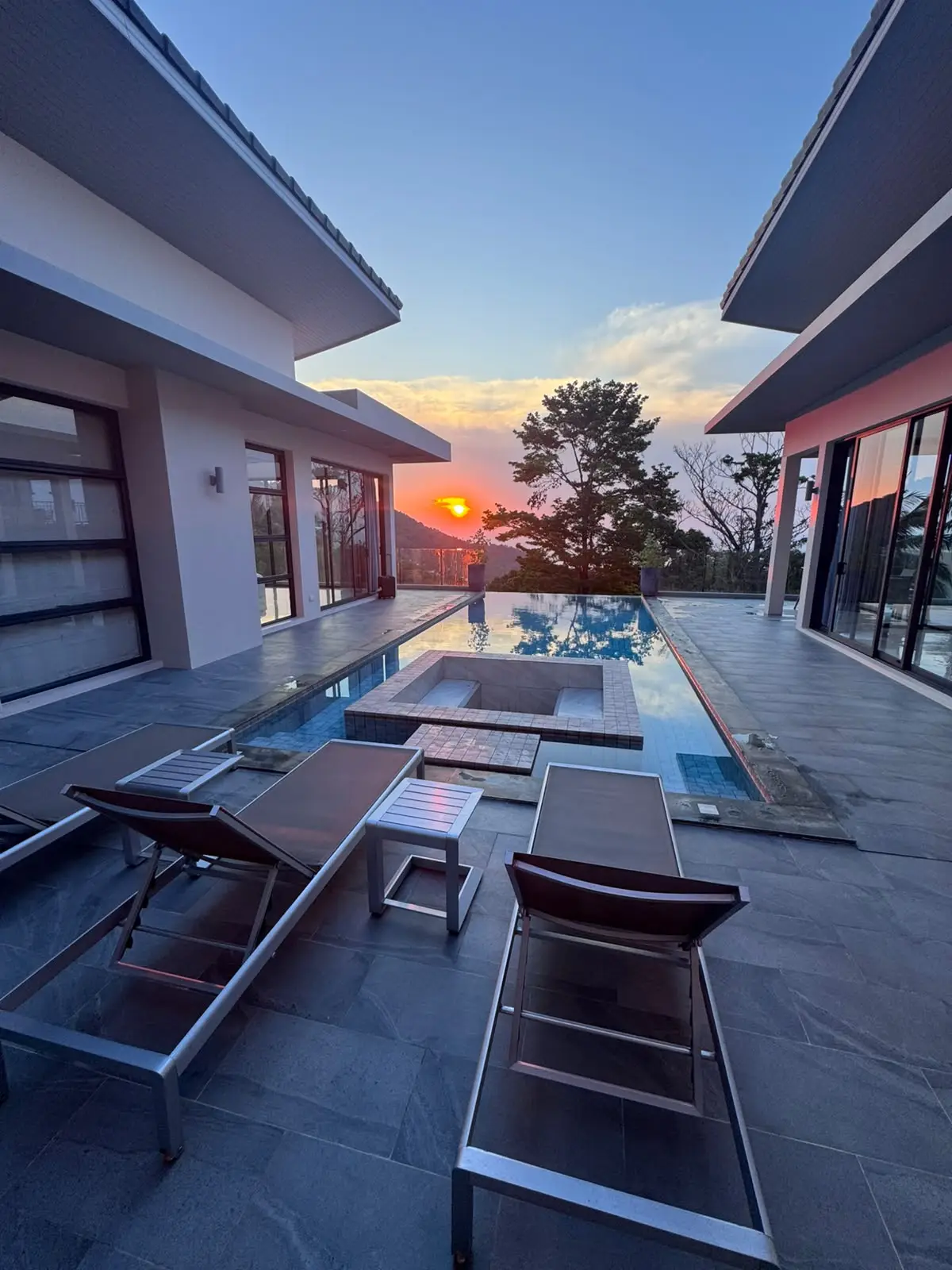 Villa contemporaine au coucher du soleil, piscine à débordement et transats sur une terrasse en pierre.