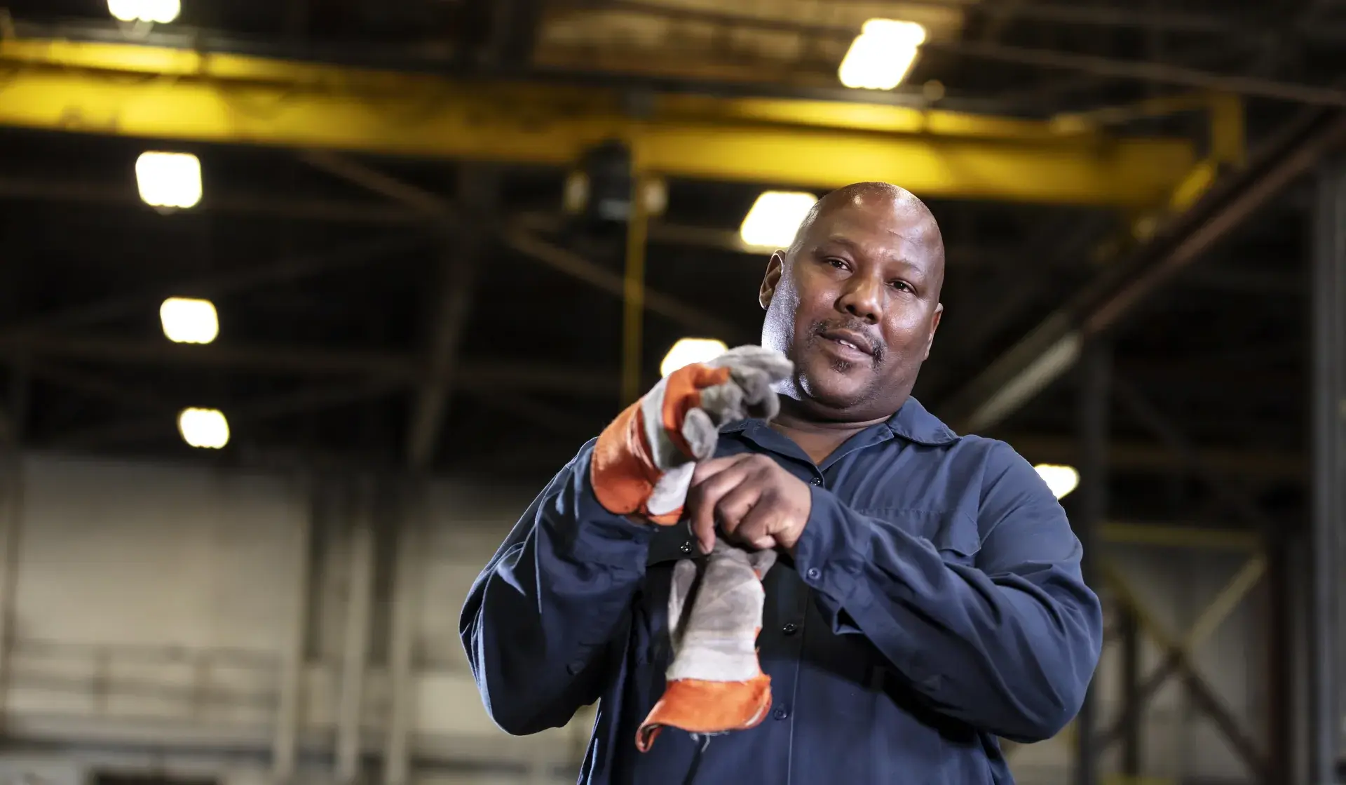 Worker in blue uniform putting on orange and gray protective gloves in an industrial setting.