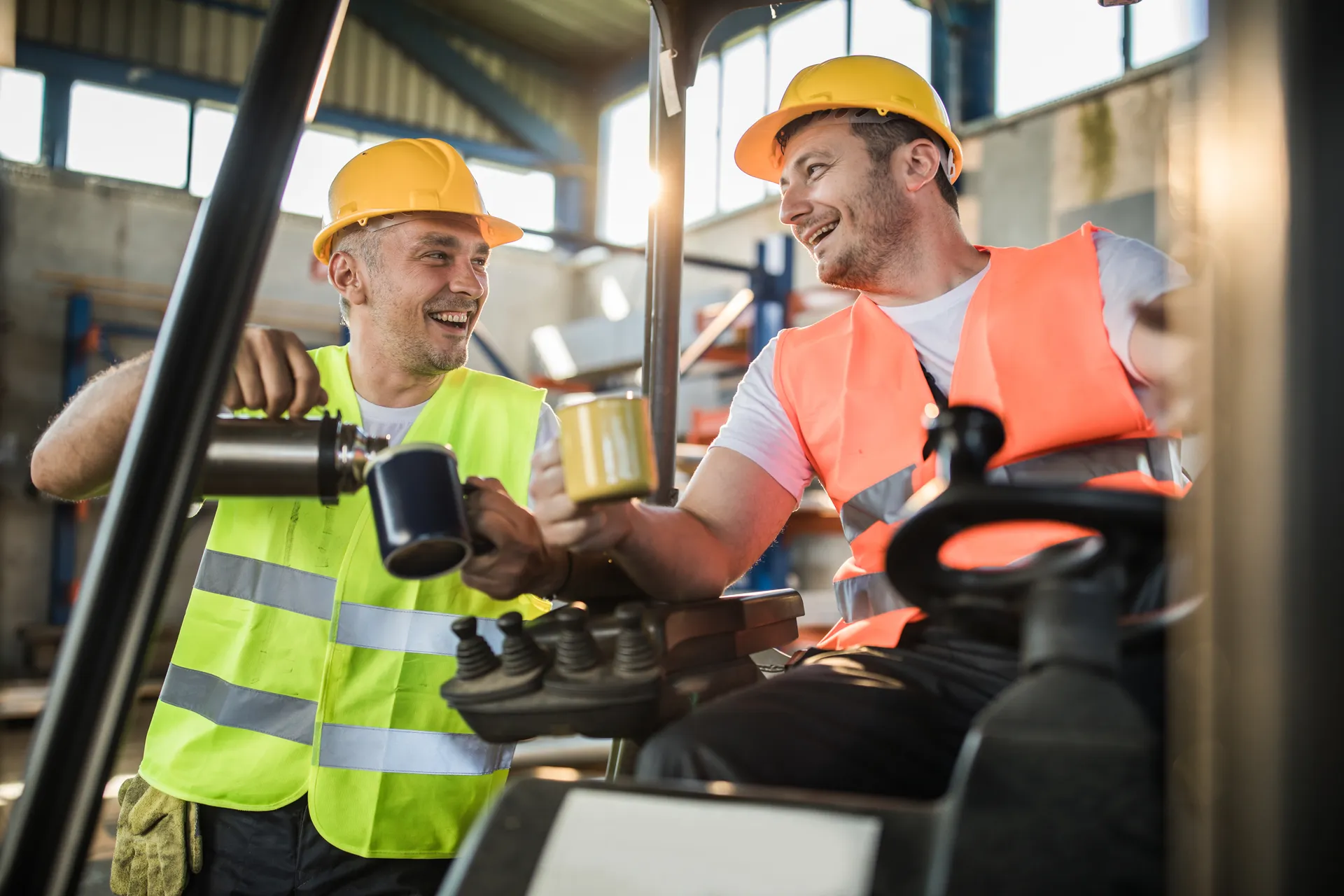 Two construction workers in helmets and vests smiling and sharing coffee in a warehouse.