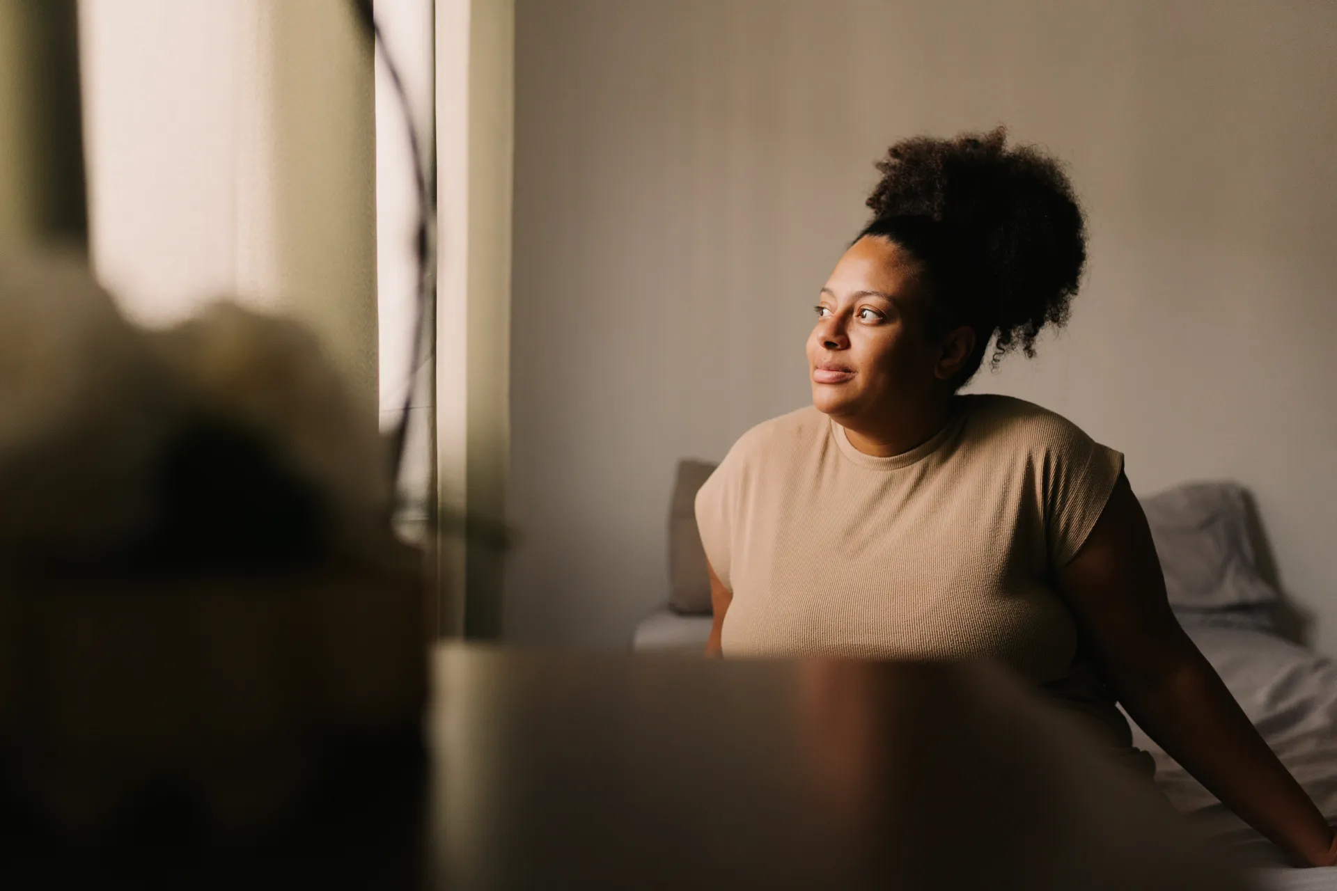 Woman with curly hair sitting on bed, looking thoughtfully out a window with soft light.