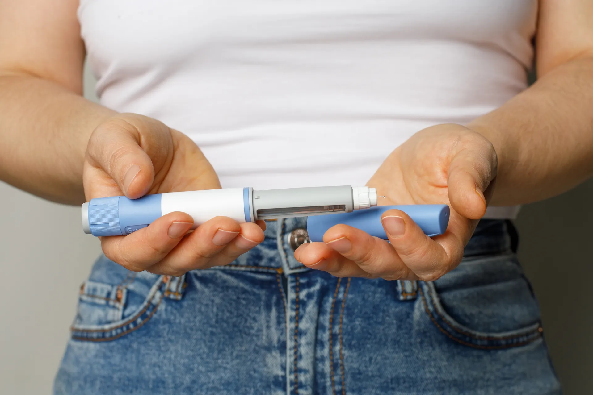 Person holding an insulin pen with needle cap removed, wearing blue jeans and a white shirt.