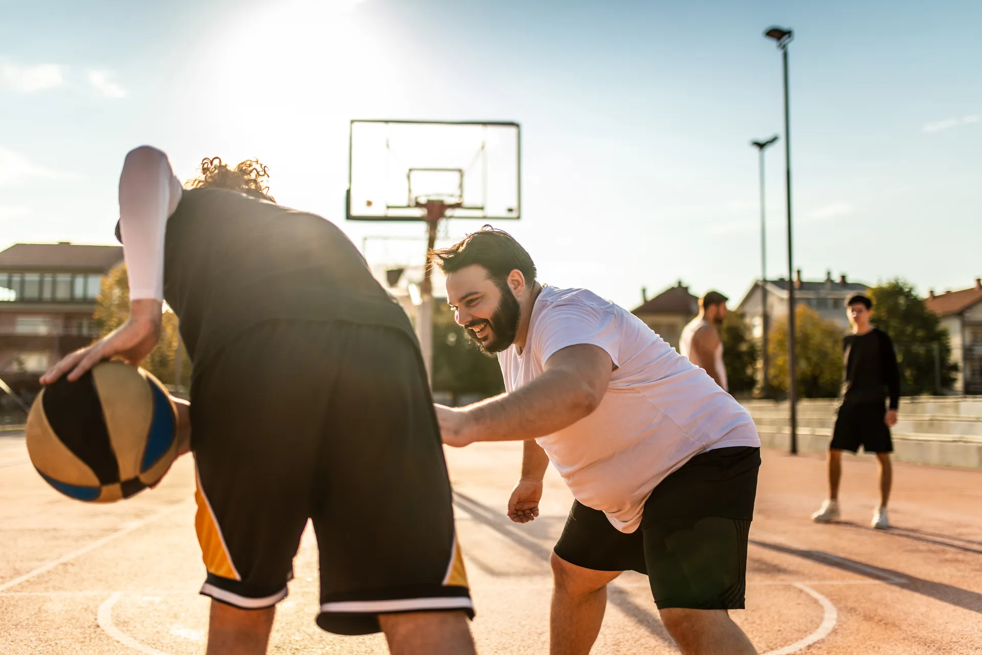 Two men playing basketball outdoors, one dribbling and the other defending closely, on a sunny day.