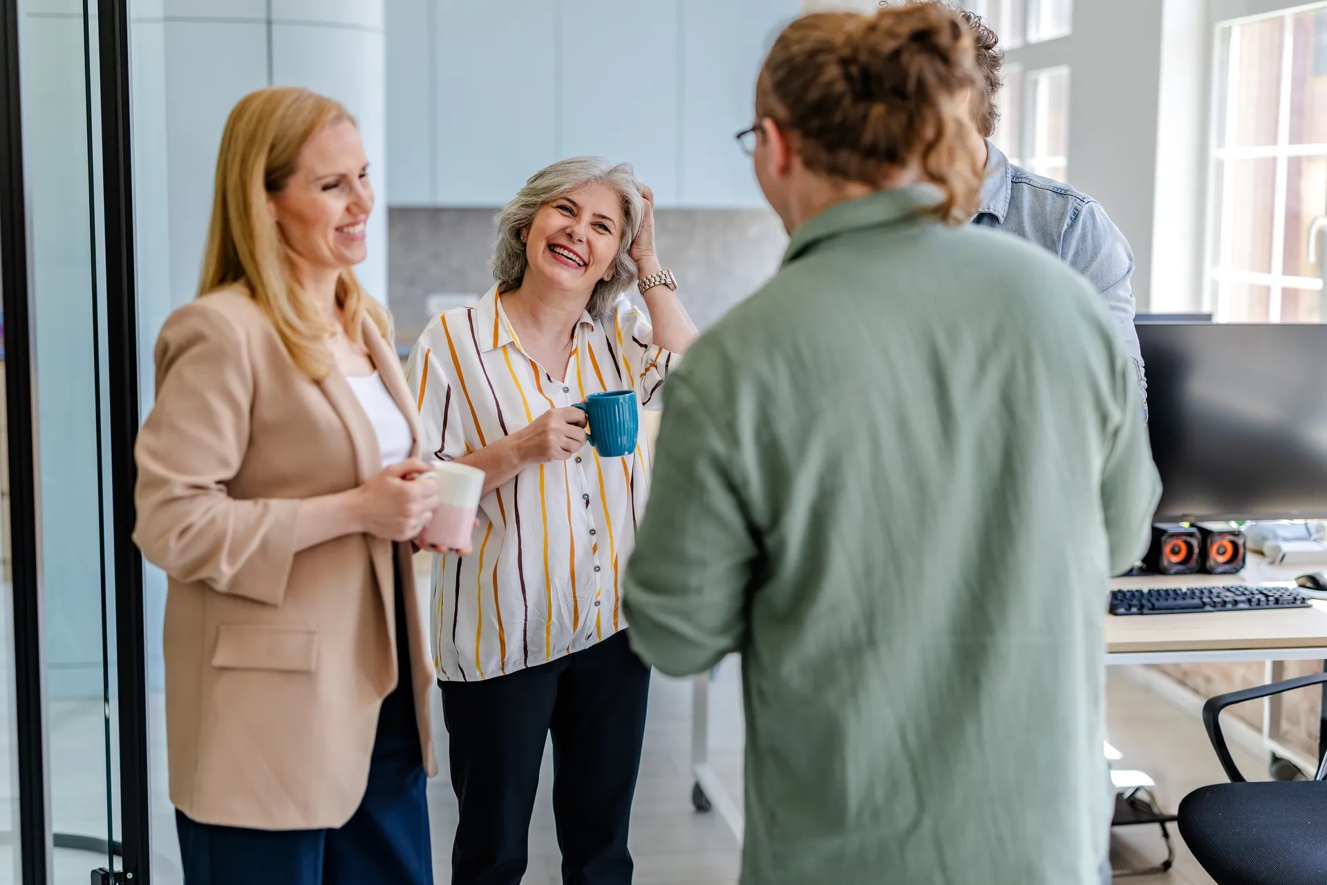 Four coworkers standing in office chatting and laughing while holding coffee mugs.