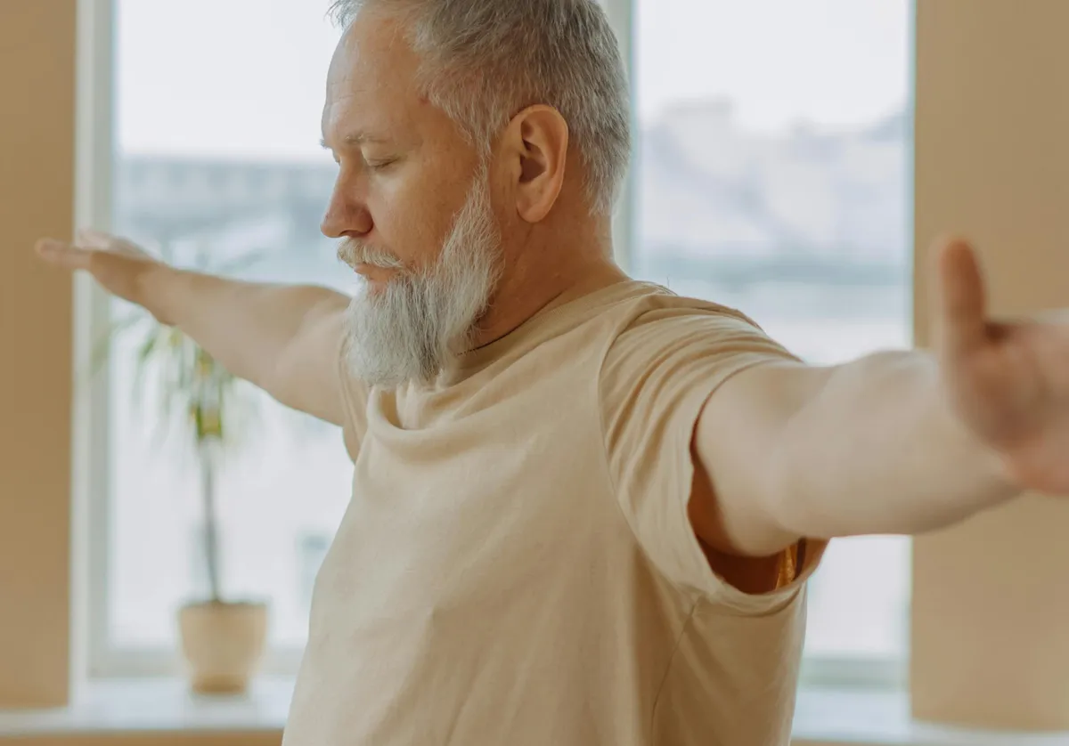 Older man with gray beard extending arms and closing eyes near a window with a potted plant.