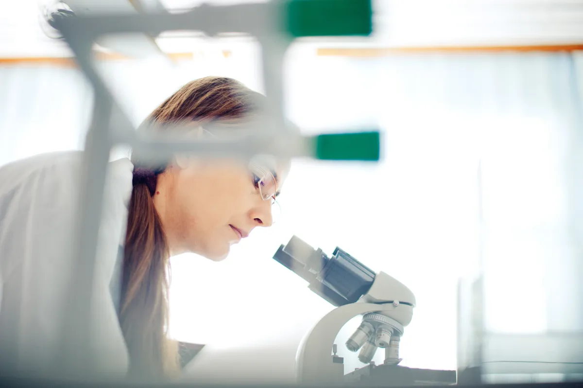 Female scientist with glasses looking through a microscope in a bright laboratory.
