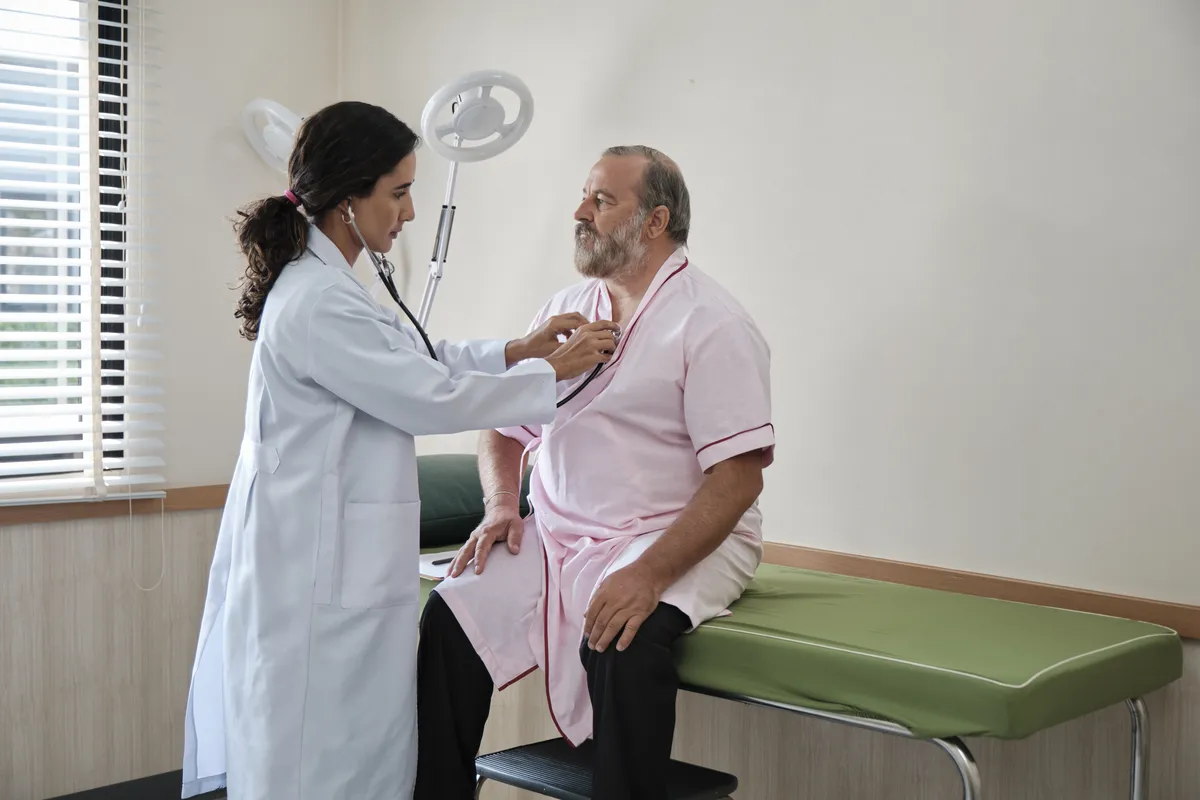 Female doctor using stethoscope to check an older man's chest while he sits on exam table.