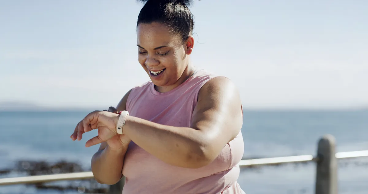 Smiling woman in pink sleeveless shirt checking fitness tracker by the sea on a sunny day.
