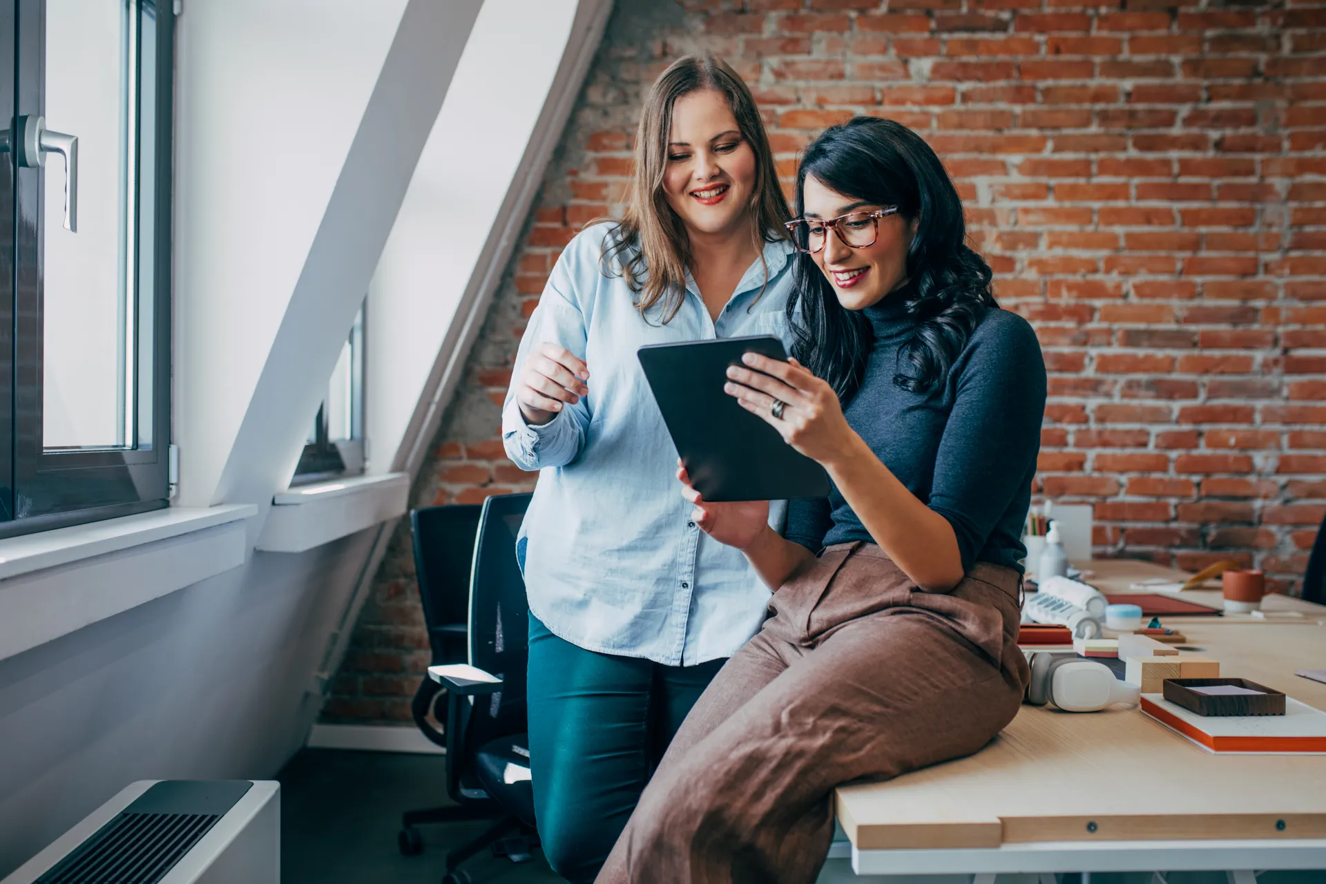 Two women smiling and looking at a tablet inside a modern office with a brick wall.