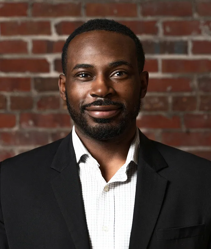 Smiling man with short beard in a black blazer and checkered shirt in front of a brick wall.