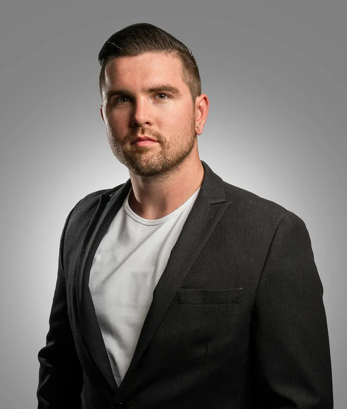 Serious man with short hair and beard wearing a black blazer and white shirt on grey background.