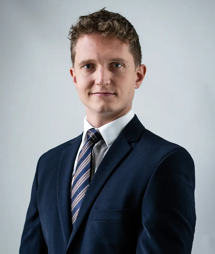 Young man with short curly hair wearing a navy suit, white shirt, and striped tie on gray background.