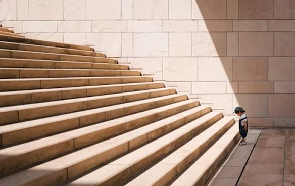 Small child stands at the bottom of large, wide stone steps against a tiled wall.