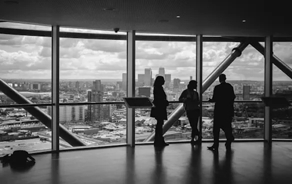 Three people silhouettes standing by large windows overlooking a cityscape with skyscrapers on a cloudy day.