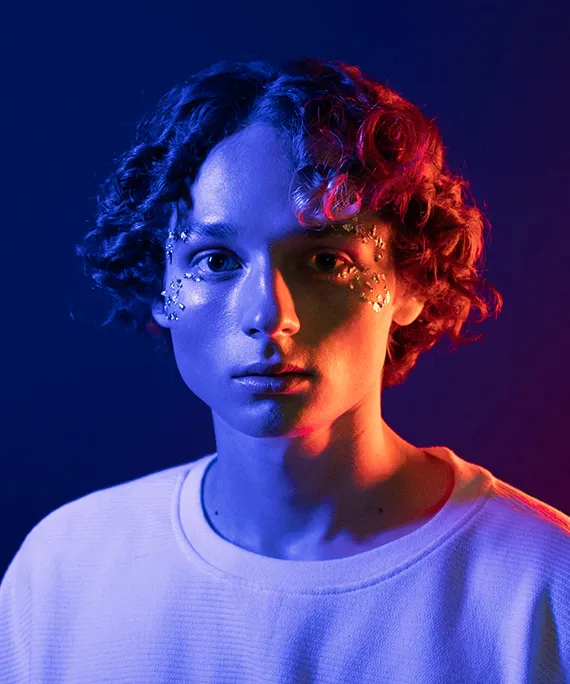 Portrait of a person with curly hair and face glitter in blue and red lighting wearing a white shirt.