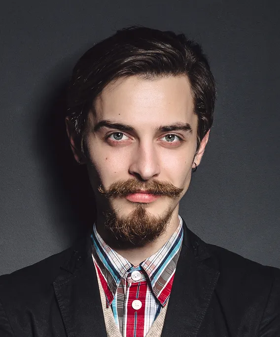 Man with styled mustache and beard wearing a plaid shirt and black blazer against a dark background.
