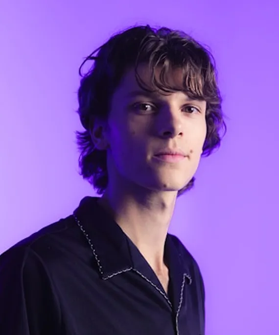 Young man with tousled brown hair wearing a black shirt, against a purple background.