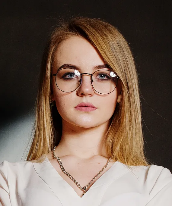 Portrait of a young woman with straight blonde hair, round glasses, and a chain necklace on dark background.