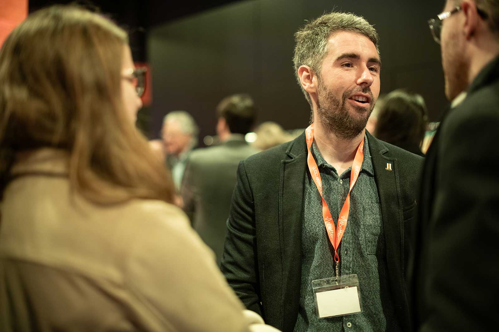 Hombre con gafete naranja conversando con otras personas en un evento social o profesional.