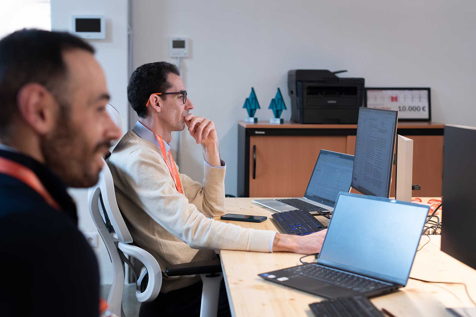 Dos hombres en una oficina trabajando frente a varias pantallas de computadora, uno con gafas y ambos con gafetes naranjas.