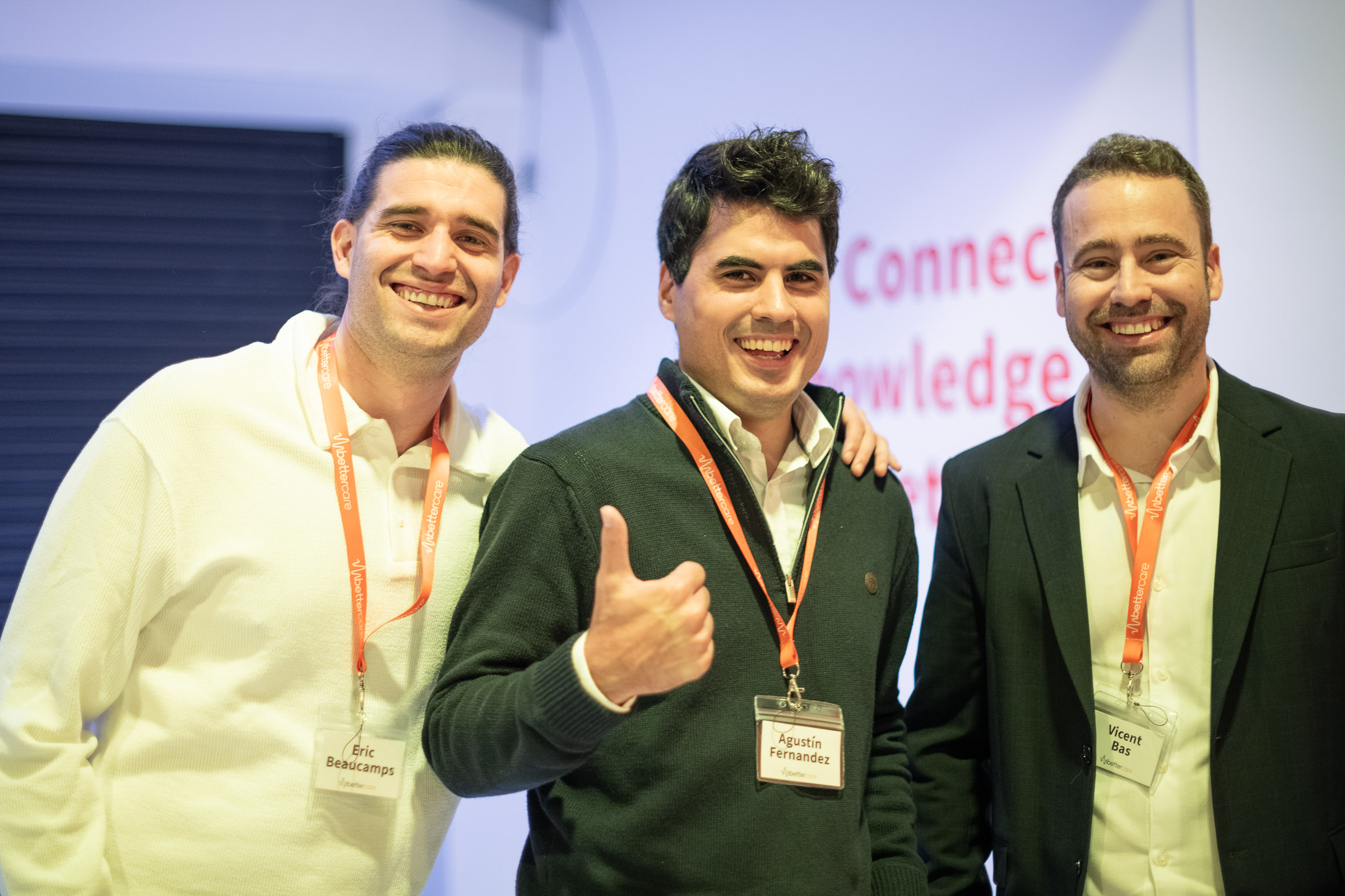 Tres hombres sonrientes con gafetes de identificación y collares naranjas en un evento corporativo.