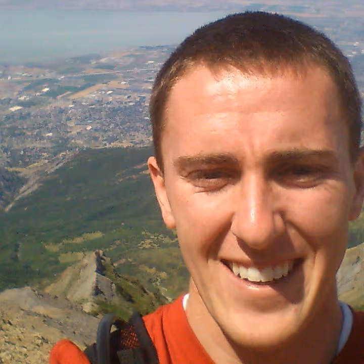 An image of a man with short hair and a backpack standing on top of a mountian