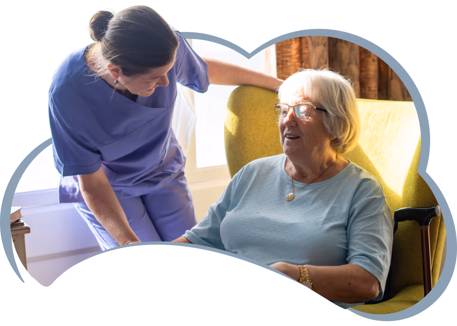 Caregiver in scrubs leaning toward smiling elderly woman seated in a yellow chair with a cane nearby.