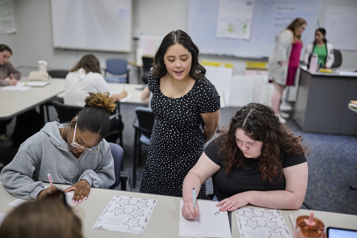 Teacher assisting two students writing at desks in a classroom with other students in background.