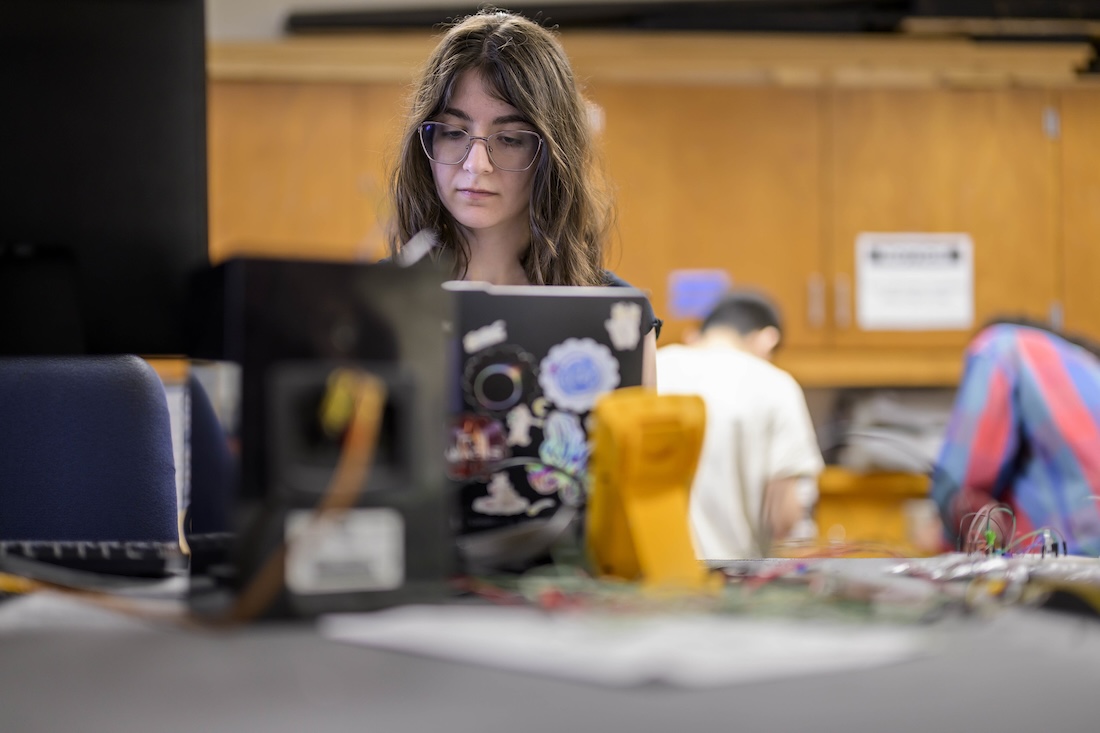 A woman with glasses is working on a laptop covered in stickers at a table indoors.